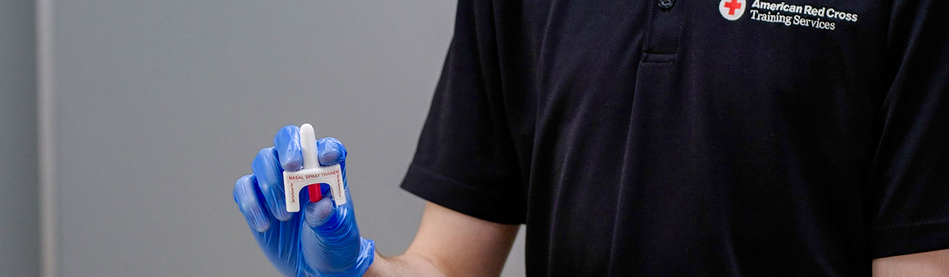 A person wearing an American Red Cross shirt holding a naloxone nasal spray training device.