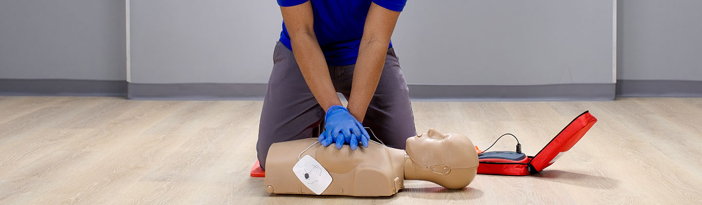 A person wearing blue gloves performing compressions on a CPR manikin that has AED pads attached with an AED trainer on the floor nearby.