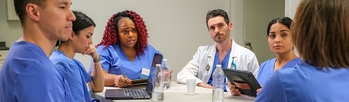 Neonatal Advanced Life Support training session with medical staff in scrubs and a doctor discussing around a table in a clinical setting.