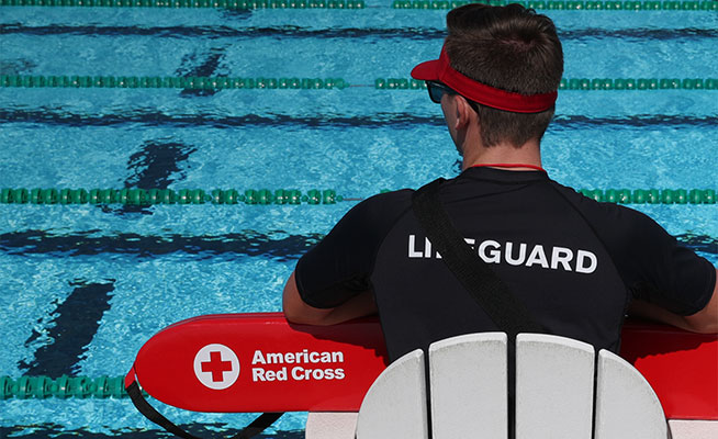A lifeguard holding a Red Cross Rescue Tube overlooking a swimming pool.