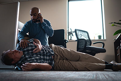 A man on the phone calls for help while tending to another injured man who is laying on the ground in distress.