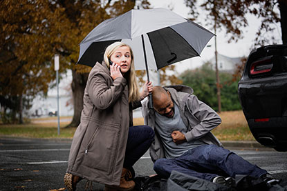 Woman holding an umbrella on a phone tending to a man in distress until help arrives.