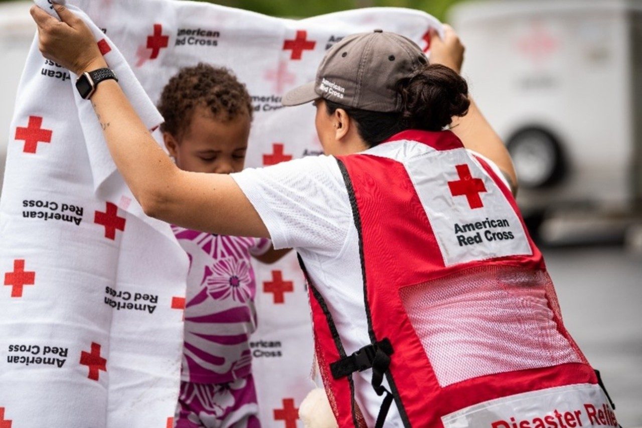 A Red Cross volunteer wearing a red Disaster Responder vest wraps a Red Cross blanket around a little boy.