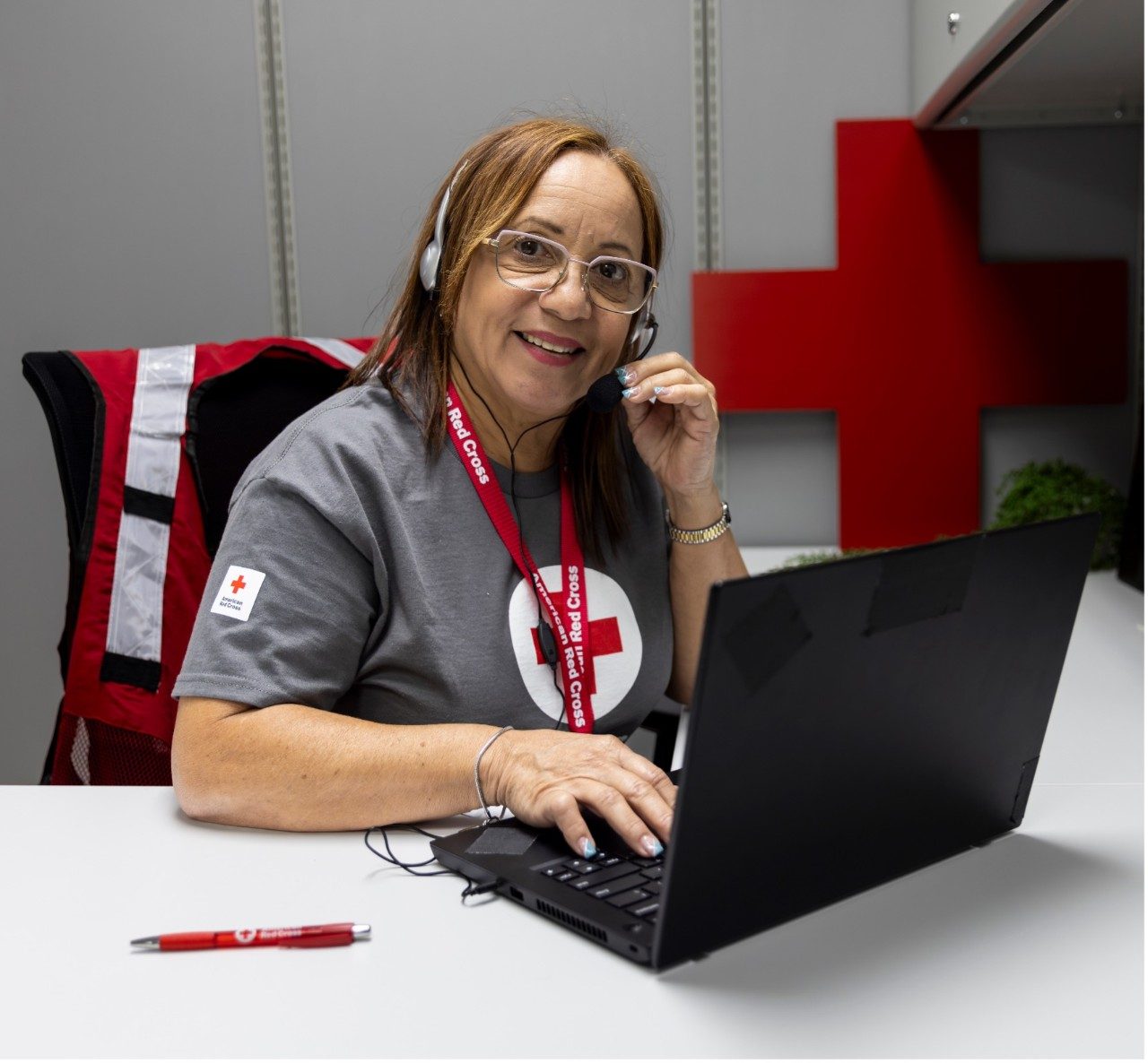 A woman with shoulder-length auburn hair sitting at a desk answering phones while wearing a Red Cross t-shirt
