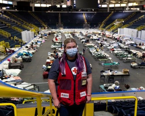 red cross volunteer wearing a mask stands on an elevated platform overlooking rows of beds in shelter behind her