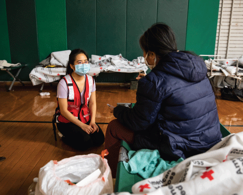 red cross volunteer wearing a mask kneeling in front of shelter resident who is sitting on a cot.