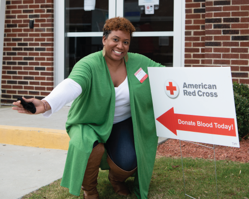 Red Cross volunteer kneeling next to a Donate Blood sign outside.