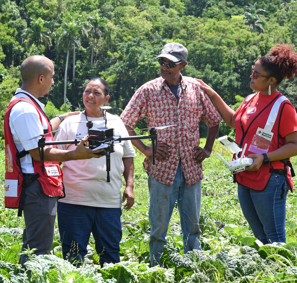 Fotografía de dos voluntarios de la Cruz Roja con dos personas, sosteniendo un dron para tomar fotos aéreas .