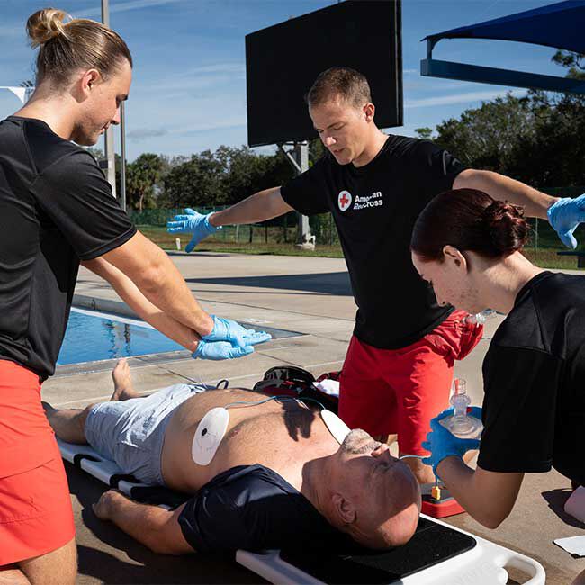 A group of lifeguards using an AED and rescue mask to resuscitate an unconscious person near a swimming pool.