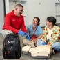 Red Cross CPR instructor talking to two students next to a CPR manikin and Red Cross Backpack.