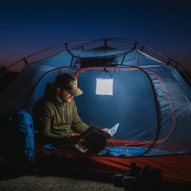 The LuminAID Max QI Lantern illuminated and hung up in a camping tent with a person reading a manual.